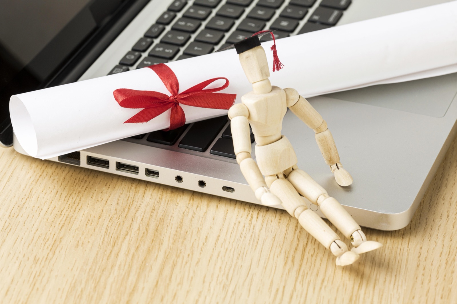 A wooden figurine in a graduation cap sitting on a laptop beside a rolled certificate tied with a red ribbon