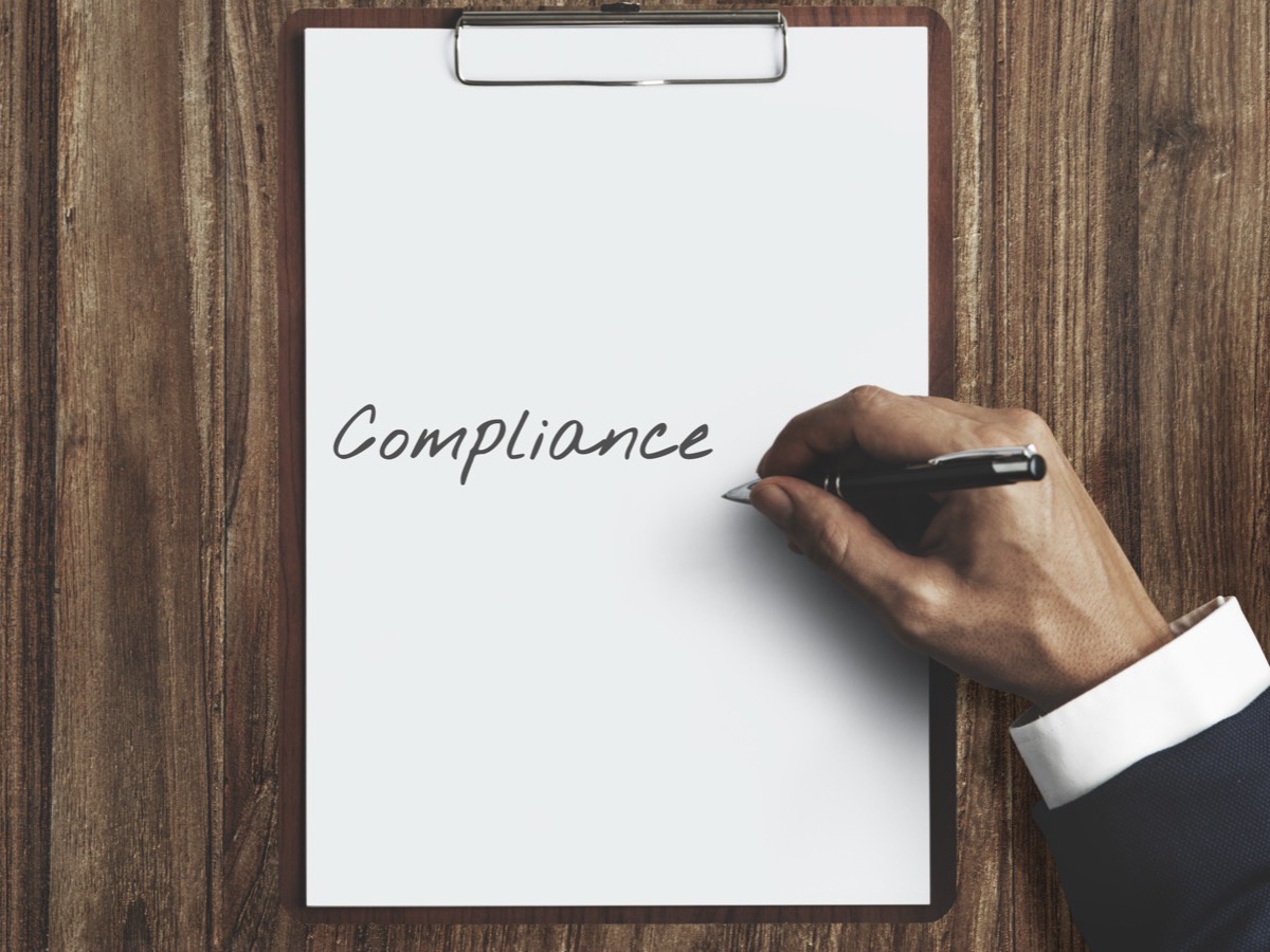 A hand writing the word Compliance on a clipboard