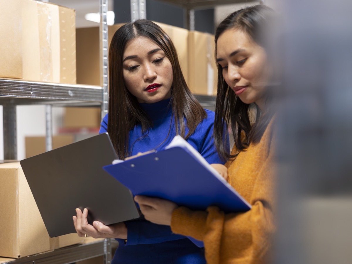 Two warehouse staff checking a procedure on a clipboard between stock shelves